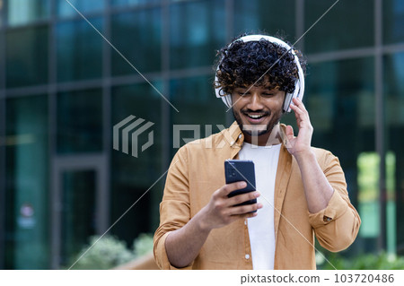 A young male Hispanic student is standing on the street near the campus wearing headphones with a phone in his hands. Listens to music, a podcast, talks on a video call. A young male Hispanic student is standing on the street near the campus wearing headphones with a phone in his hands. Listens to music, a podcast, talks on a video call. 103720486