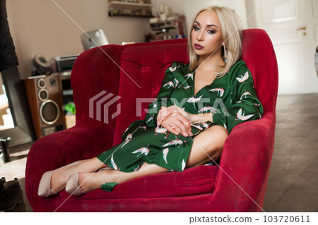 Gorgeous blonde woman with makeup in green dress sitting on red sofa 103720611