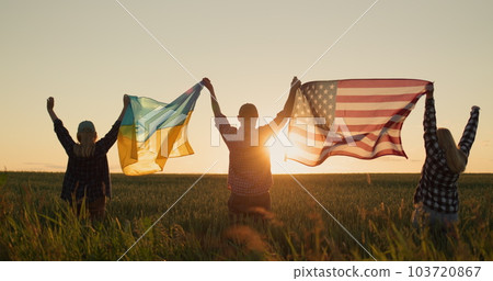 A group of friends raises the flag of the United States and the flag of Ukraine over their heads. Standing against the backdrop of a field of wheat where the sun sets 103720867