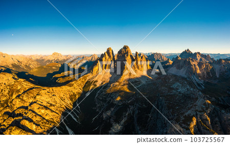 Rocky peaks of Tre Cime di Lavaredo at sunrise. Mountain landscape of wide hilly canyons and valley with curved roads under clear sky aerial view 103725567