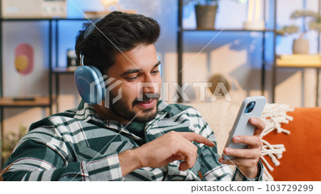 Happy indian man in wireless headphones relaxing sitting on sofa at home listening favorite music Happy indian man in wireless headphones relaxing sitting on sofa at home listening favorite music 103729299