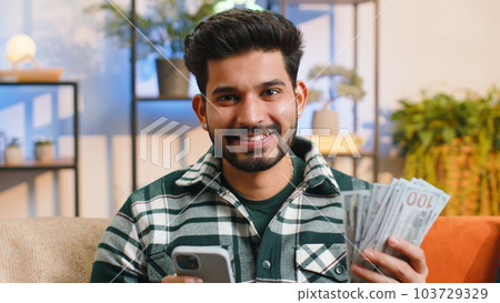 Smiling happy indian man counting money cash use smartphone, income saves lottery win budget at home 103729329