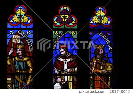 Colorful medieval era stained glass panels depicting monarchs, saints set in stone gothic tracery in a cathedral. Illuminated windows against a dark background. Blues, greens, reds 103740043