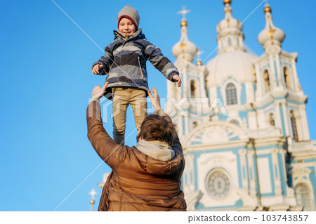 Young father throws up his cute and little son in the air in front of cathedral. Father's Day Concept game with child Young father throws up his cute and little son in the air in front of cathedral. Father's Day Concept game with child 103743857