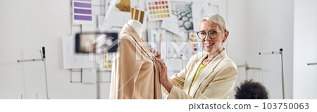 Smiling woman tailor shows new beige jacket shooting video while African-American seamstress sits at workplace 103750063