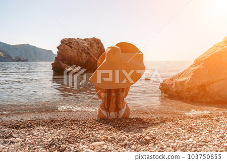 woman sea travel Tanned middle-aged woman with long hair and a white bathing suit. He sits on the seashore in a large sun hat with his back and looks at the sea. woman sea travel Tanned middle-aged woman with long hair and a white bathing suit. He sits on the seashore in a large sun hat with his back and looks at the sea. 103750855