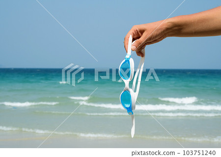 Hand holds swimming goggles agaist seascape. Tropical vacation concept. 103751240