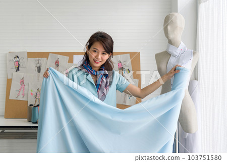 Young asian dressmaker in striped scarf check the quality of blue fine fabric from the roll. Fashion designer room. Young asian dressmaker in striped scarf check the quality of blue fine fabric from the roll. Fashion designer room. 103751580
