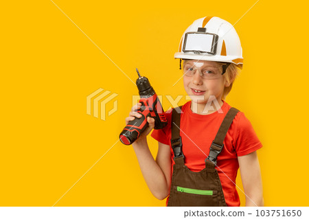 Portrait of boy as worker on yellow background. Child wears protective helmet and holds drill in his hand. Copy space, mock up 103751650
