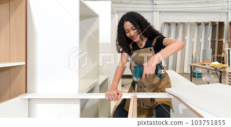 Teenager in brown apron use  cordless electric drill with battery to assemble wooden furniture in the factory. Wooden cabinets of various sizes are placed in the work area. 103751855