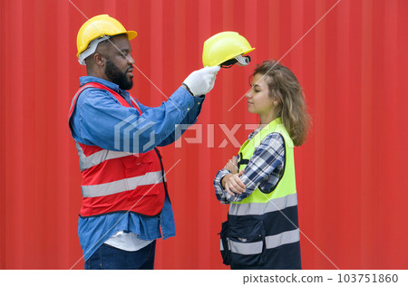A foreman with mustache and beard helps a female apprentice wear a hardhat. Both of them wearing reflective safety vest. A large cargo container is in the background. A foreman with mustache and beard helps a female apprentice wear a hardhat. Both of them wearing reflective safety vest. A large cargo container is in the background. 103751860