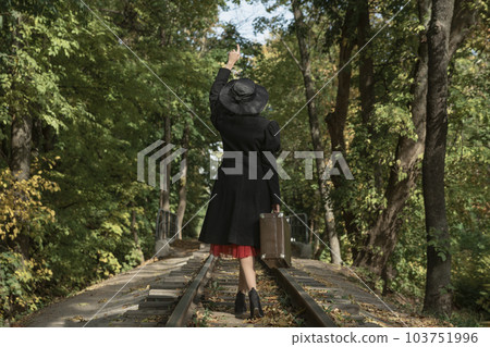 Woman in stylish coat, broad-brim hat and with vintage suitcase walks along railway tracks going through forest. Back view 103751996