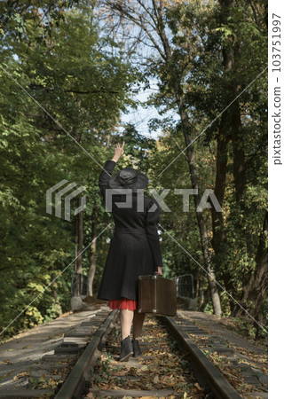 Stylish woman in coat, broad-brim hat and with suitcase walks along railway tracks going through forest. Back view Stylish woman in coat, broad-brim hat and with suitcase walks along railway tracks going through forest. Back view 103751997