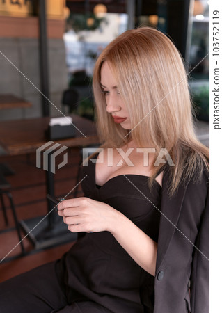 Portrait of blonde girl in black bustier outfit. Young woman wears top with deep neckline sitting in cafe. Vertical frame. 103752119
