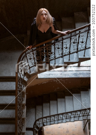 Beautiful blonde girl in black outfit with deep neckline stands on an old staircase. Top view. Vertical frame. 103752122