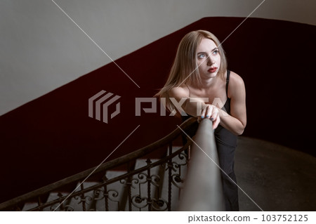 Young sad fair-haired woman near stairs in blouse with deep neckline. Model poses for photo indoor. 103752125