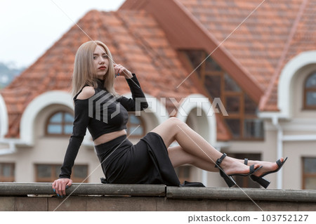 Beautiful young woman with blonde hair wears black clothes long skirt and heels. Stylish girl outside. Old building on background Beautiful young woman with blonde hair wears black clothes long skirt and heels. Stylish girl outside. Old building on background 103752127