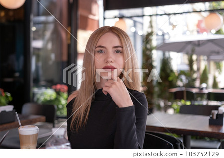 Portrait of young girl with blonde hair in cafe. Attractive young fair-haired woman sits at table in cafe and looks pensively into the camera. 103752129