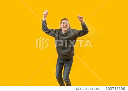 Teenage boy screams or laughs with hands above his head and jumping. Isolated against yellow background. Copy space. Teenage boy screams or laughs with hands above his head and jumping. Isolated against yellow background. Copy space. 103752308