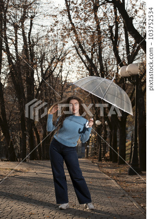 Pretty young woman dancing and fun with transparent umbrella. Happy young woman with long hair in autumn park. Pretty young woman dancing and fun with transparent umbrella. Happy young woman with long hair in autumn park. 103752534