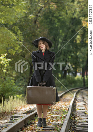 Woman in black hat and coat holds retro valise. Madame in clothes of 30-40s style mysteriously. Vertical frame 103752535