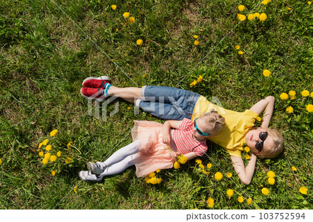 Little boy and his younger sister are lying on green field with dandelions. Siblings rest in the grass. Top view Little boy and his younger sister are lying on green field with dandelions. Siblings rest in the grass. Top view 103752594