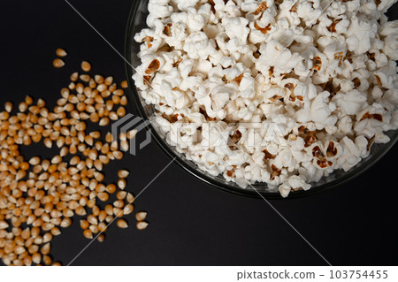Ready-to-eat popcorn in a glass bowl and corn kernels on a black background. 103754455