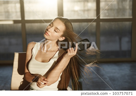 Young woman sitting on a chair and brushing her hair with a comb. Brunette woman sitting in a bright studio with big windows. Girl wearing white t-shirt. Young woman sitting on a chair and brushing her hair with a comb. Brunette woman sitting in a bright studio with big windows. Girl wearing white t-shirt. 103757071