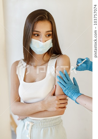 Young woman wearing mask and receiving coronavirus vaccine. A young girl receiving an immunity shot in the forearm. Closeup of doctor in blue gloves giving Covid-19, AIDS or flu antivirus vaccine to Young woman wearing mask and receiving coronavirus vaccine. A young girl receiving an immunity shot in the forearm. Closeup of doctor in blue gloves giving Covid-19, AIDS or flu antivirus vaccine to 103757074