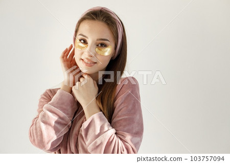 Young woman applying a face patches. Brunette woman standing on a white backgroung. Girl wearing rose pajamas and headband. Young woman applying a face patches. Brunette woman standing on a white backgroung. Girl wearing rose pajamas and headband. 103757094