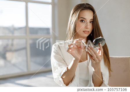Young woman sitting on a chair and applying mascara. Brunette woman sitting in a bright studio with big windows. Girl wearing white blouse. Young woman sitting on a chair and applying mascara. Brunette woman sitting in a bright studio with big windows. Girl wearing white blouse. 103757106