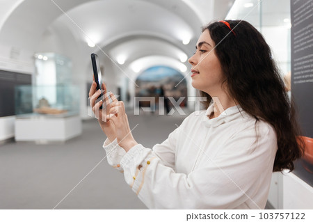 Side view of young Caucasian woman using smartphone. Concept of cultural exhibition and modern educational excursion 103757122