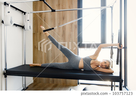 Pregnant woman doing exercises with a special equipment. Fit blonde woman working out in a gym. Woman wearing grey sportive leggins and top. 103757123