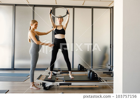 Two young pregnant women doing exercises on a special equipment. Fit women working out in a gym. Women wearing grey and black sportive leggins and tops. 103757156