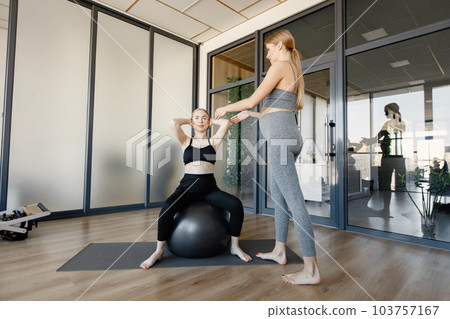 Two pregnant women doing exercises with a finess ball. Fit blonde and brunette woman working out in a gym. Women wearing grey and black sportive leggins and tops. 103757167