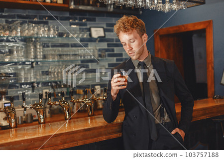 Man in a black suit drinks alcohol. Attractive guy drinks whiskey from a glass.. 103757188