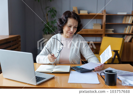 Middle aged senior woman sit with laptop and paper document. Smiling older mature lady reading paper bill pay online at home managing bank finances calculating taxes planning loan debt pension payment Middle aged senior woman sit with laptop and paper document. Smiling older mature lady reading paper bill pay online at home managing bank finances calculating taxes planning loan debt pension payment 103757688