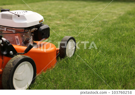 Gardening work tools. Close up details of orange electric lawn mower, wheels, motor on bright lush green grass. Rotary lawn mower machine cut lawn. Professional lawn care service. Place for text 103760258