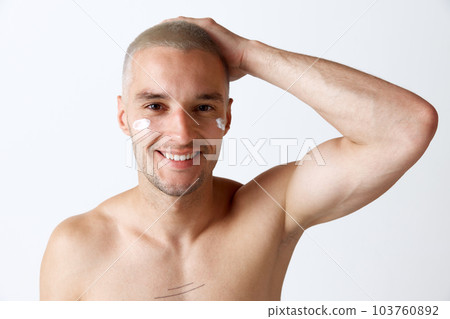 Portrait of young handsome smiling man taking care after skin with moisturizing cream against white studio background 103760892