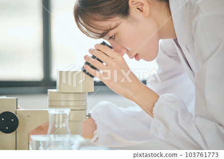 A young woman in a white coat looking into a microscope 103761773