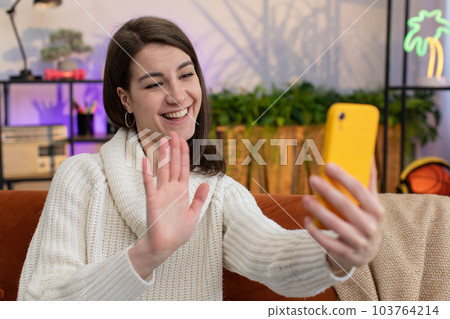 Young woman girl making online selfie video call talking waving hand hello looking at camera at home Young woman girl making online selfie video call talking waving hand hello looking at camera at home 103764214
