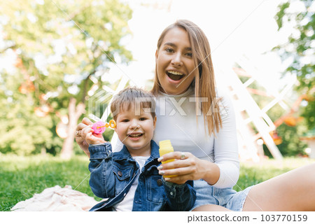 Happy and cheerful mom and son are blowing soap bubbles in a summer park, sitting on the grass 103770159