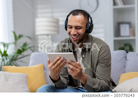 Young smiling hispanic man sitting on sofa at home wearing headphones and using tablet. Young smiling hispanic man sitting on sofa at home wearing headphones and using tablet. 103770243