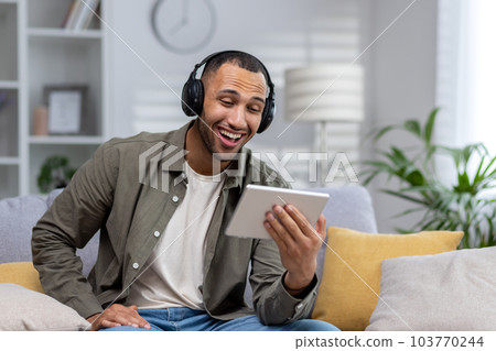 African american smiling young man sitting on couch at home wearing headphones and holding tablet, talking on phone call, watching video. African american smiling young man sitting on couch at home wearing headphones and holding tablet, talking on phone call, watching video. 103770244