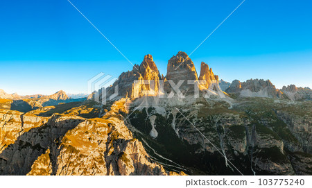 Sun rises over mountain range. High rocky peaks with sand-covered foothills of Three Peaks of Lavaredo under cloudless sky aerial view in back lit 103775240