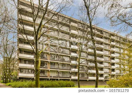 an apartment building with trees in the fore, and some cars parked on the other side of the parking lot 103781270