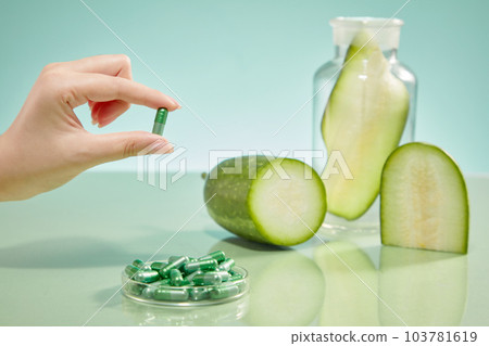 Front view of a hand holding a capsule, a petri dish containing capsules and fresh slices of winter melon on color background. Scene for advertising cosmetic product with winter melon ingredient. Front view of a hand holding a capsule, a petri dish containing capsules and fresh slices of winter melon on color background. Scene for advertising cosmetic product with winter melon ingredient. 103781619