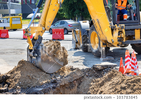 The bucket of a large construction excavator digs a trench on the road against the backdrop of a city street on a sunny summer day. 103783845