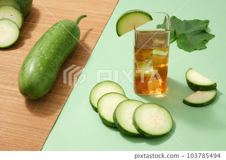 Advertising scene for drinking product with ingredient from winter melon. A cool glass cup of tea decorated with fresh winter melon, slices and leaf on green background. Advertising scene for drinking product with ingredient from winter melon. A cool glass cup of tea decorated with fresh winter melon, slices and leaf on green background. 103785494