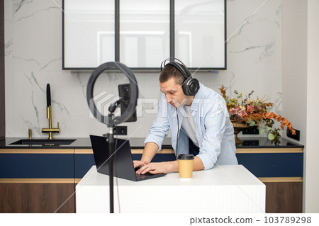 A young man conducts a video broadcast on the phone. Work in a laptop with headphones 103789298